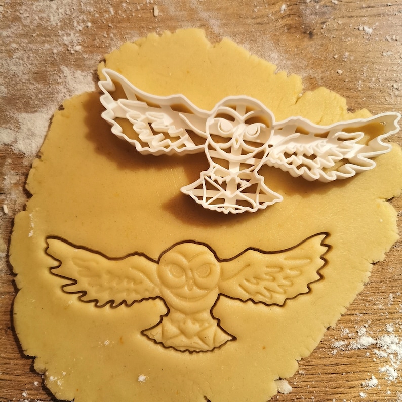 May include: A white plastic owl cookie cutter on top of a brown cookie dough with an owl shape cut out of it.