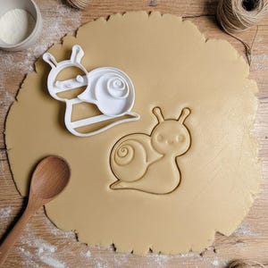Könnte beinhalten: Ein weißer, schneckenförmiger Ausstecher neben einem schneckenförmigen Abdruck im Teig. Ein Holzlöffel und eine Schüssel mit weißem Pulver befinden sich im Hintergrund. Der Teig liegt auf einer Holzoberfläche. Eine Schnurkugel befindet sich in der oberen rechten Ecke.