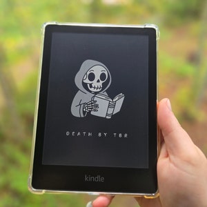 May include: A black and white illustration of a skeleton wearing a hooded robe and reading a book. The text "DEATH BY TBR" is displayed below the image. The image is displayed on a Kindle e-reader.
