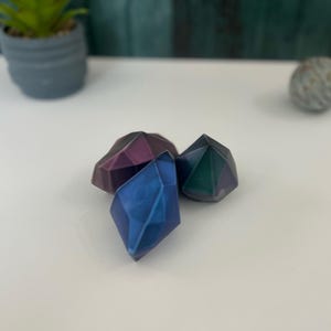 Puede incluir: Tres formas geom&eacute;tricas iridiscentes en tonos de morado, azul y verde. Las formas recuerdan a gemas y tienen un dise&ntilde;o facetado.