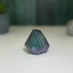 Puede incluir: Un pisapapeles geom&eacute;trico en forma de diamante, de color verde oscuro y morado. El pisapapeles tiene un acabado brillante e iridiscente.