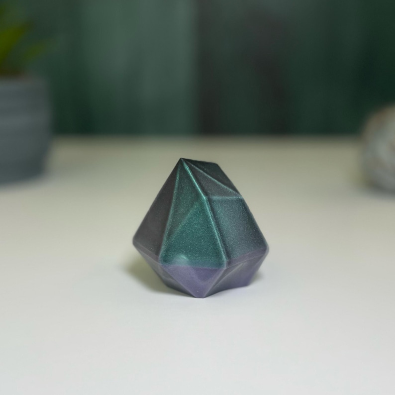 Puede incluir: Un pisapapeles geom&eacute;trico en forma de diamante, de color verde oscuro y morado. El pisapapeles tiene un acabado brillante e iridiscente.