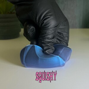 Puede incluir: Un juguete blando azul y negro con forma de roca con la palabra "SQUISHY" impresa en letras rosas.