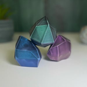 Puede incluir: Tres objetos geom&eacute;tricos, facetados, con forma de piedra preciosa. El m&aacute;s grande es de color verde oscuro con una base negra, el del medio es de color azul oscuro con una base negra, y el m&aacute;s peque&ntilde;o es de color morado con una base negra.
