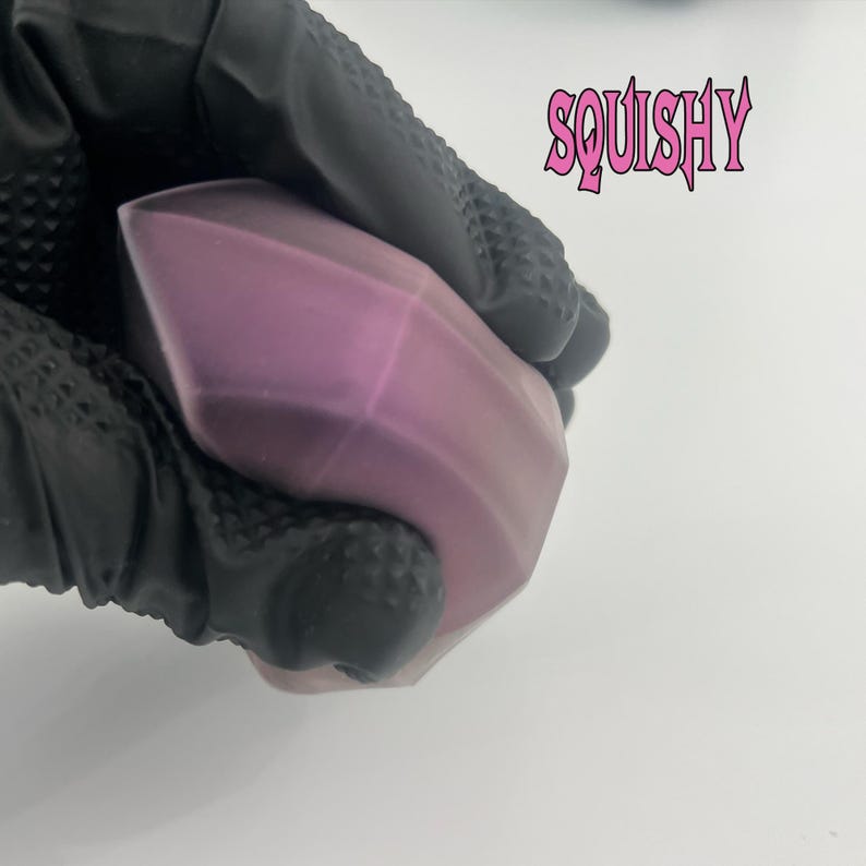 Puede incluir: Un juguete blando de color morado y rosa con forma de prisma geom&eacute;trico. El juguete est&aacute; siendo sostenido por una mano enguantada de negro. La palabra "SQUISHY" est&aacute; escrita en letras rosas sobre un fondo blanco.