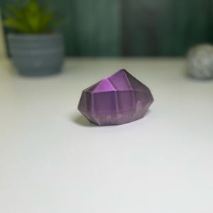 Puede incluir: Un cristal geom&eacute;trico morado con forma de diamante. El cristal tiene una superficie lisa y pulida.