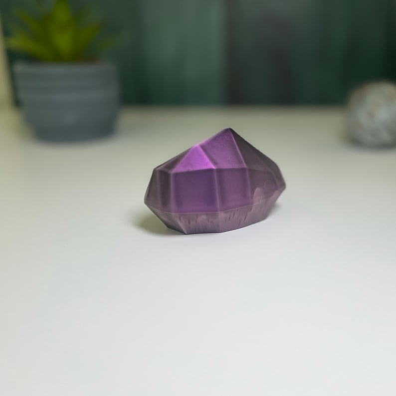 Puede incluir: Un cristal geom&eacute;trico morado con forma de diamante. El cristal tiene una superficie lisa y pulida.