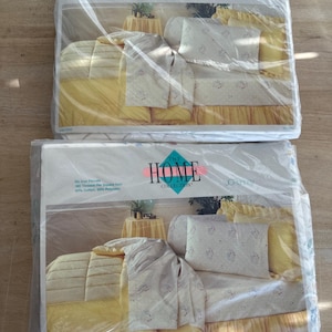 May include: Two packages of The Home Collection bed linens in a floral print. The packaging shows a yellow bed with a white floral patterned bedspread and pillowcases. The text on the packaging reads "The Home Collection" and "No Iron Percale 180 Threads Per Square Inch 50% Cotton, 50% Polyester."