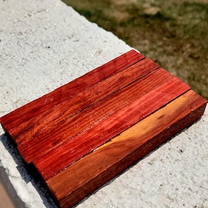 May include: A stack of six wooden dowels with a reddish-brown color and a natural wood grain. The dowels are arranged in a staggered pattern, with the top dowel slightly offset from the bottom dowel.