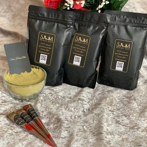 May include: Three black pouches of henna powder with gold lettering that reads "SAM HENNA ARTS" and a QR code. The pouches are on a white, textured surface with three brown henna cones and a bowl of henna powder.