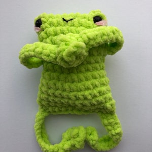 May include: A lime green crocheted frog plush toy with black eyes and pink cheeks. The frog is sitting up with its arms crossed and its legs curled up.