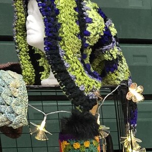 May include: A handmade crochet hood in shades of green, black, and purple, displayed on a mannequin head. The hood features a long, pointed design and a black pom-pom. A mug cosy and other crochet items are also visible.