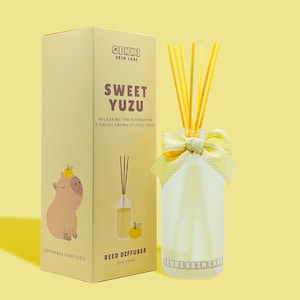 May include: A yellow box with a cartoon capybara and the text "Sweet Yuzu Reed Diffuser" and "GENKI SKIN CARE". The box is next to a clear glass bottle with a yellow bow and yellow sticks. The bottle is filled with a clear liquid.