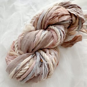 May include: A bundle of mixed fabric ribbons and rope in shades of pink, beige, and grey. The textures include woven, frayed, and twisted elements, creating a soft, tactile appearance. A small gold-coloured bead is visible.