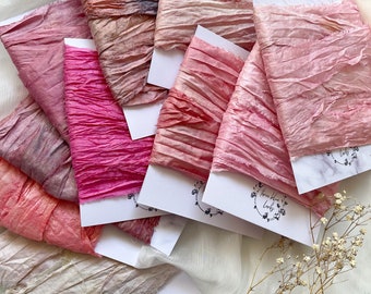 May include: A collection of hand-dyed silk ribbons in various shades of pink, peach, and mauve. The ribbons are crinkled and textured, presented on white cards with a floral logo. The arrangement is set against a soft, white fabric background.