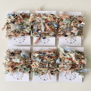 May include: Six spools of colourful yarn with a white label that says "Three Blue Larks". The yarn is a mix of textures and colours, including pink, blue, brown, and white.