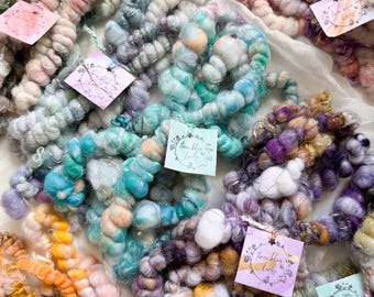 May include: A collection of colorful, chunky yarn skeins in various pastel shades, including pink, blue, green, and purple. Each skein is adorned with a small tag. The yarn appears to be hand-spun or art yarn, suitable for fiber art projects.