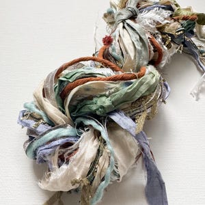 May include: A bundle of colourful silk ribbon scraps in shades of blue, green, white, and orange. The ribbon is tied together in a knot and is ready to be used for crafting projects.