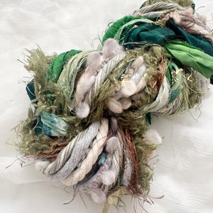 May include: A close-up of a bundle of textured yarn in shades of green, beige, and brown. The yarn appears to be a mix of materials, including thick rope-like strands, fluffy textures, and ribbons. The background is a soft, white fabric.