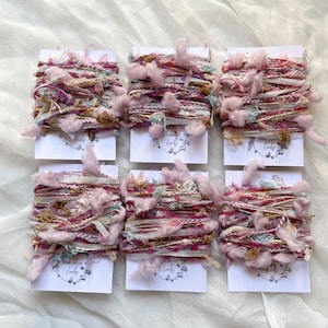 May include: Six spools of pink, white and brown yarn with a frayed texture. Each spool is wrapped in white paper with a circular logo that says "Three Blue Birds".