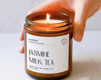 May include: A brown glass candle jar with a white label that reads "scentfield Handmade Vegan Candle Jasmine Milk Tea Creamy Vanilla / Milk / Jasmine | 9.0 oz Made in Texas". The candle is lit and the flame is visible.