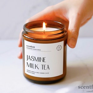 May include: A brown glass candle jar with a white label that reads "scentfield Handmade Vegan Candle Jasmine Milk Tea Creamy Vanilla / Milk / Jasmine | 9.0 oz Made in Texas". The candle is lit and the flame is visible.