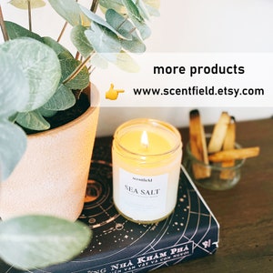 May include: A white candle with a black label that reads "scentfield SEA SALT" is lit and sitting on a black and white book with a gold star pattern. The book is titled "NH&Agrave; KH&Aacute;M PH&Aacute;" in Vietnamese.