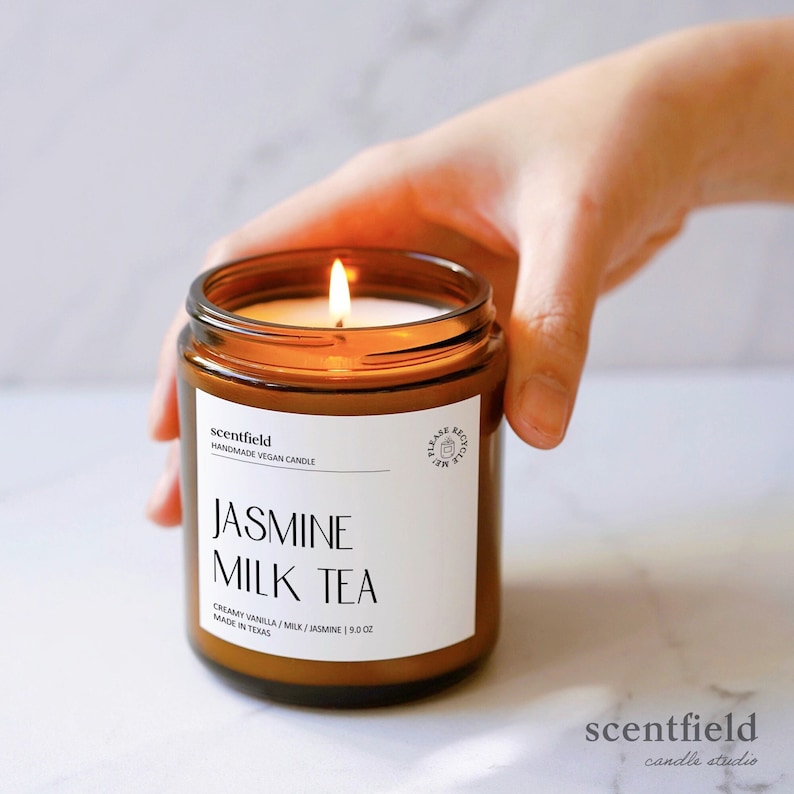 May include: A brown glass candle jar with a white label that reads "scentfield Handmade Vegan Candle Jasmine Milk Tea Creamy Vanilla / Milk / Jasmine | 9.0 oz Made in Texas". The candle is lit and the flame is visible.