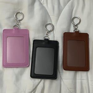 May include: Three ID badge holders in pink, black, and brown. Each holder has a clear window and a silver keyring attached. The pink holder is on the left, the black in the middle, and the brown on the right.