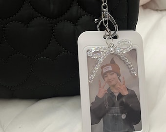 May include: A black quilted bag with heart-shaped stitching, a silver keychain, and a white ID holder. The ID holder features a photo of a person making a peace sign and a decorative bow.