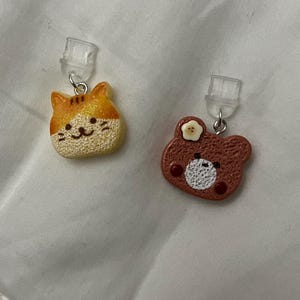 May include: A pair of clip-on earrings featuring a cat and a bear design. The cat earring is orange and tan with a smiling face. The bear earring is brown with a white circle and a small flower. Both earrings have silver-colored metal loops and clear plastic clips.