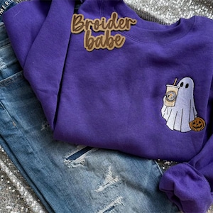 May include: A purple sweatshirt with the text "Broider babe" and an embroidered ghost holding a coffee cup and a pumpkin. The sweatshirt is paired with blue denim jeans.