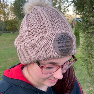 Op de afbeelding: Een roze gebreide beanie met een nep bont pompon en een bruin leren patch met de tekst "mind your own motherhood".