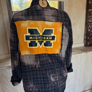 Könnte beinhalten: Ein blau-graues Flanellhemd mit Karomuster und einem großen gelben Aufnäher auf dem Rücken. Der Aufnäher zeigt das Wort "MICHIGAN" in marineblauen Großbuchstaben. Das Hemd hat ein verwaschenes, verblichenes Aussehen.