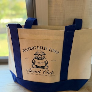 May include: A canvas tote bag with navy blue accents. The bag features a front pocket with the text "Foxtrot Delta Tango Social Club" and a graphic of a bear reading a book. The bag has navy blue handles and a navy blue base.