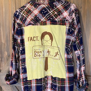May include: A blue and red plaid flannel shirt with a yellow patch sewn on the back. The patch features a cartoon character with glasses and the text "FACT. scion Fall Retreat 2019 Rule #1 Don't Die".