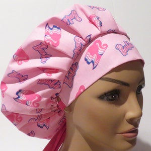 May include: Pink scrub cap with a repeating pattern of the word "Barbie" in a script font and a stylized graphic. The cap is made of a soft fabric and has a gathered design for a comfortable fit.