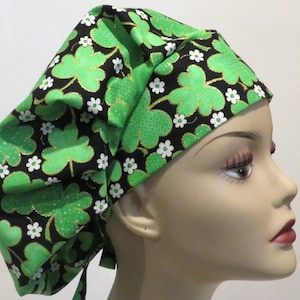 Puede incluir: Un gorro de quirófano verde y negro con un patrón de trébol y flores blancas. El gorro tiene un lazo en la parte posterior.
