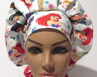 May include: White scrub cap featuring colorful illustrations of animated princesses and floral accents. The cap has a gathered design and red ribbon ties. The fabric is patterned with images of Ariel, Jasmine, and other characters.