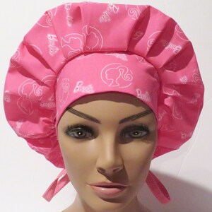 May include: Pink surgical scrub hat with a white Barbie logo pattern. The hat has a gathered top and ties at the back.
