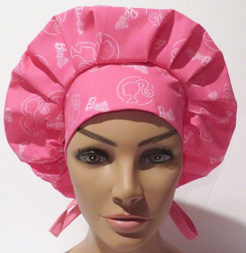 May include: Pink surgical scrub hat with a white Barbie logo pattern. The hat has a gathered top and ties at the back.