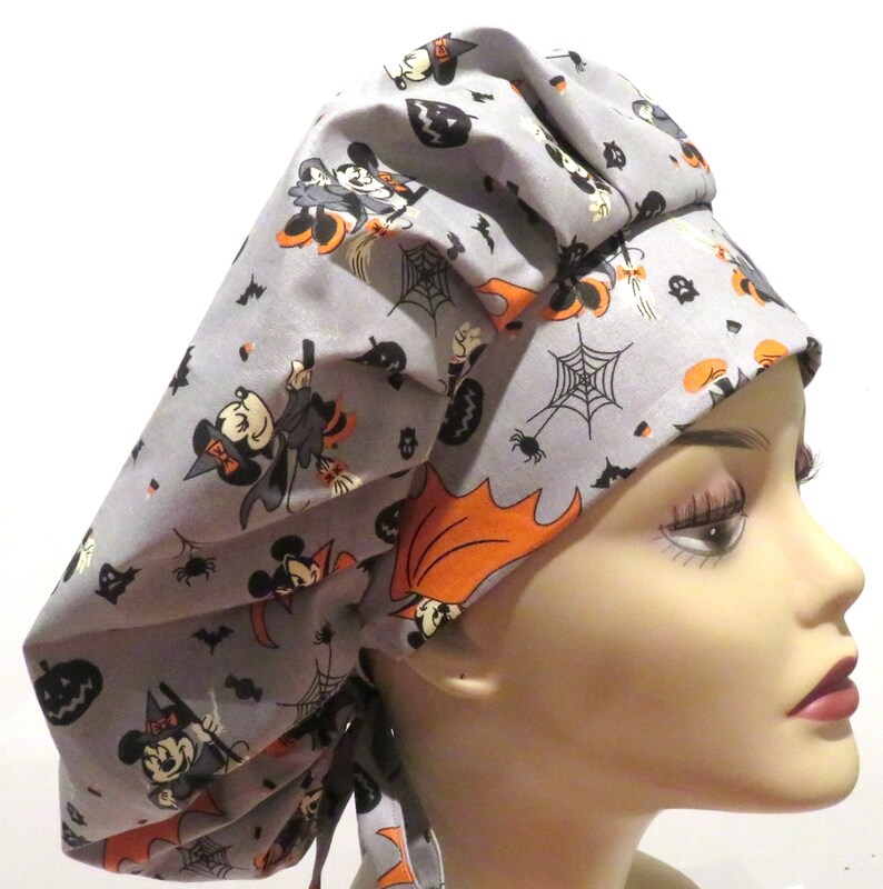 May include: A gray scrub hat with a Halloween-themed print featuring Mickey Mouse and Minnie Mouse dressed as witches. The print also includes bats, pumpkins, and spiderwebs.