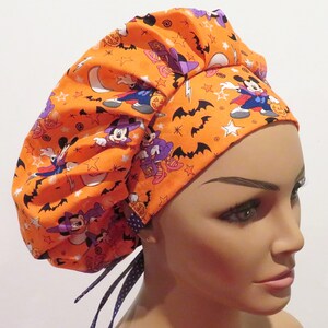 Puede incluir:  gorro de quirófano naranja con un estampado de Mickey Mouse de Halloween con murciélagos, estrellas y lunas. El gorro tiene detalles en morado y azul y lazos.