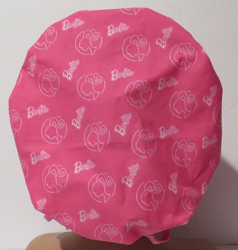 May include: Pink fabric shower cap with a white outline of a woman's face and the word "Barbie" repeated throughout.
