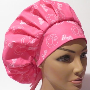 May include: Pink surgical scrub hat with a white Barbie logo pattern. The hat has a gathered top and ties at the back.