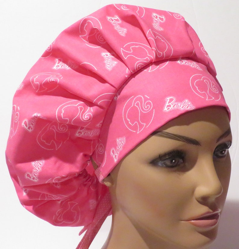 May include: Pink surgical scrub hat with a white Barbie logo pattern. The hat has a gathered top and ties at the back.