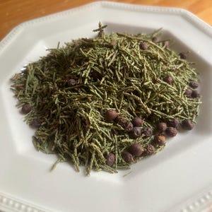 May include: A white, scalloped-edge plate holds a pile of dried green juniper needles and dark brown juniper berries. The arrangement is centered on the plate, with the berries scattered among the needles.