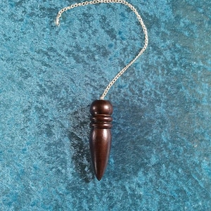 May include: A dark brown wooden pendulum with a silver chain, resting on a teal velvet surface. The pendulum has a conical shape with a rounded top and is designed for divination and spiritual practices.