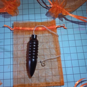 May include: A dark brown wooden pendulum with a silver chain and a small clear crystal. The pendulum is displayed inside an orange mesh bag with a matching ribbon drawstring. Several other orange bags are visible on a blue grid background.