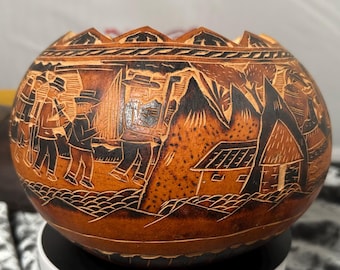 May include: A round, tan-colored gourd bowl featuring intricate, dark-toned carvings of figures in hats walking through a landscape with small houses and mountains. The top edge of the bowl has a decorative, scalloped, and geometric border design.
